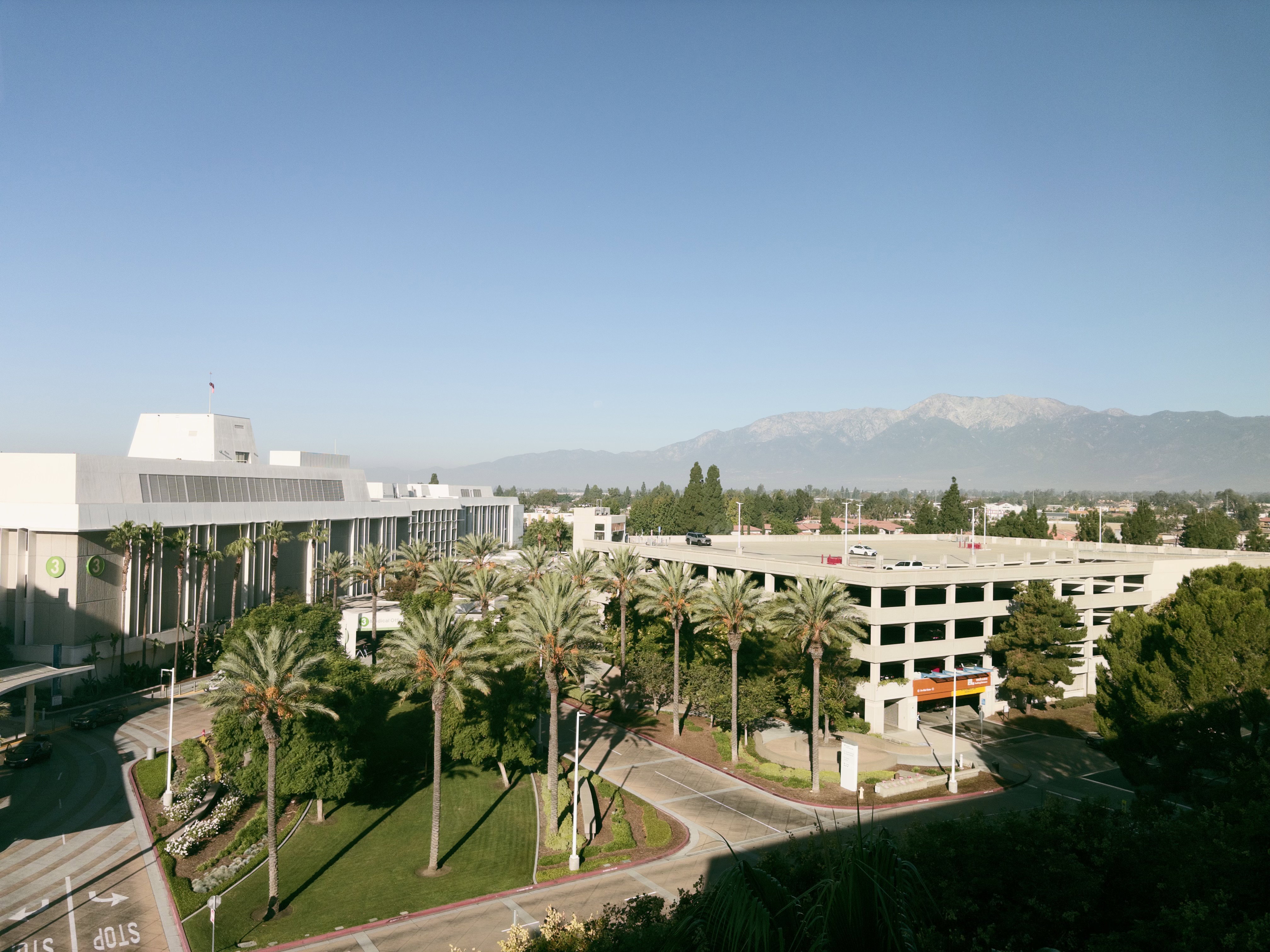A building, palm trees, and a parking structure, shot from a high vantage point, with mountains in the background, against a blue sky.