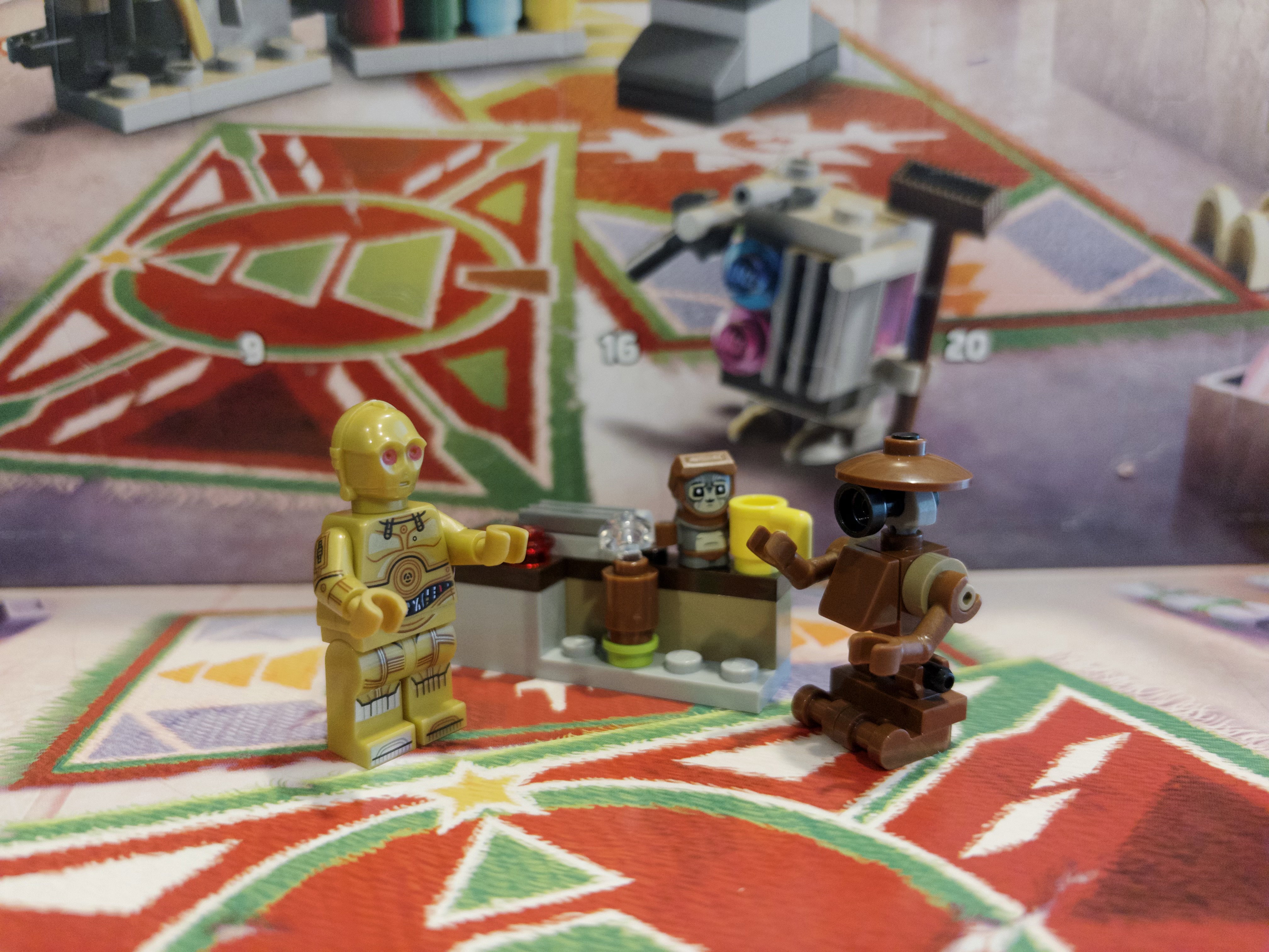 A tiny Lego figure of the tiny Star Wars character Babu Frik on a small Lego workbench with a small yellow Lego coffee mug. A C-3PO Minifig with red eyes faces off against a Lego pit droid.