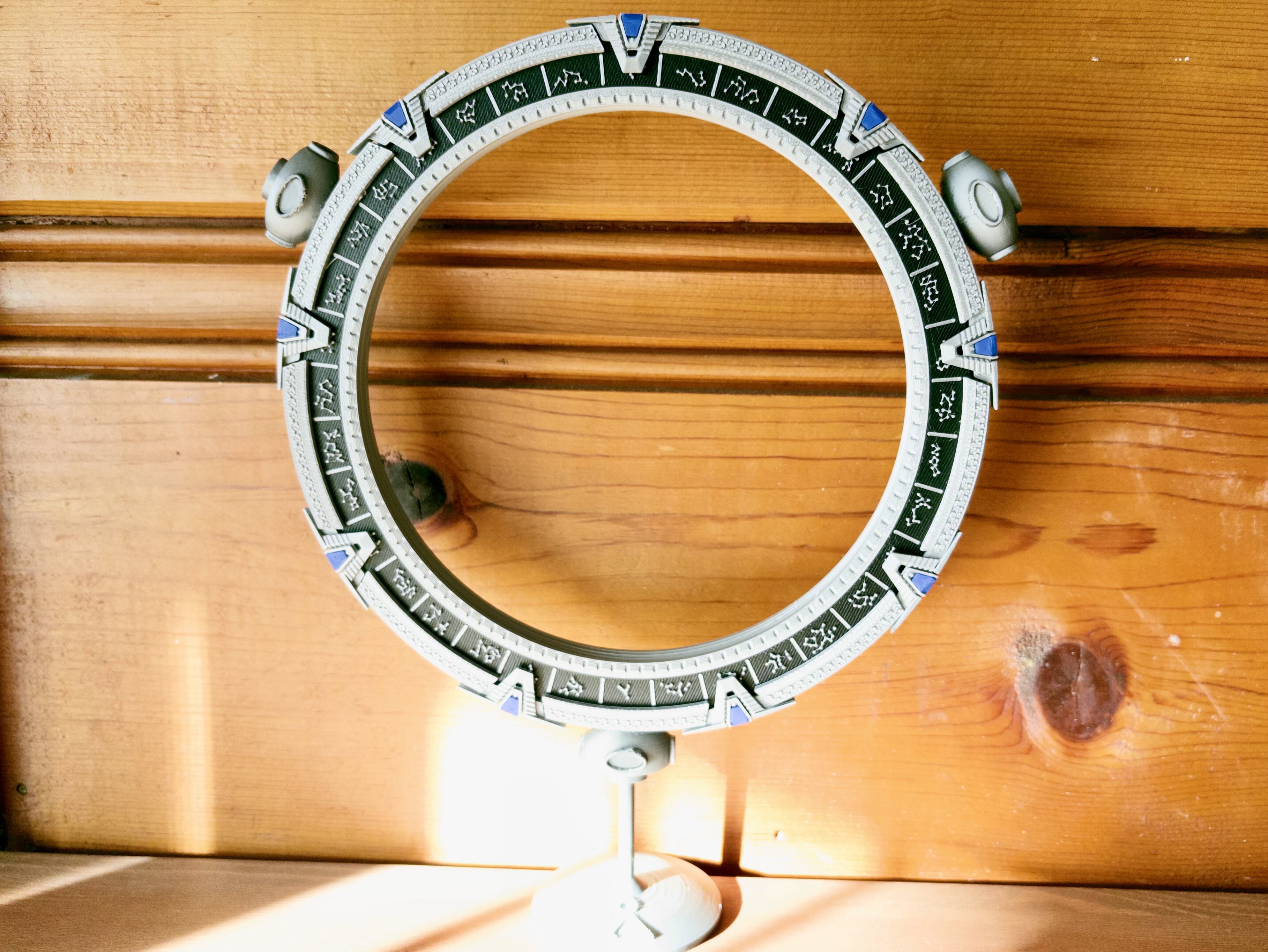 A silver, black, and blue Atargate ring hovers on a silver stand on a wood table against a wood wall.
