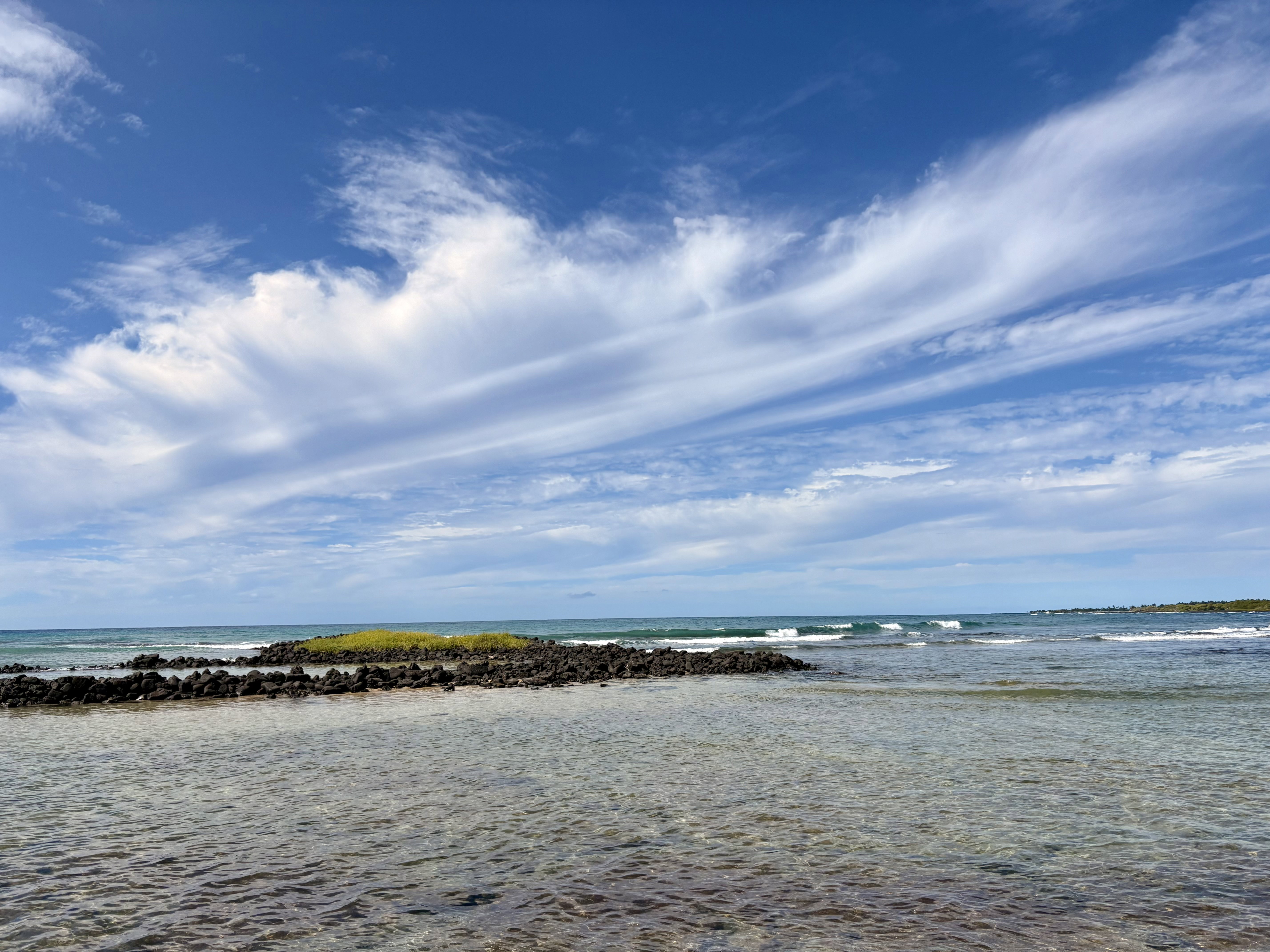 Calm beach water surrounds an area with black lava rock and grass, waves crash beyond and wispy clouds are against a blue sky.