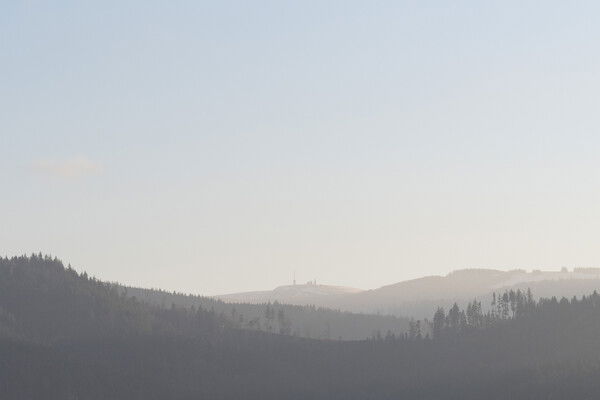 Landscape photograph taken from the Dreisamtal looking towards the Feldberg in the Black Forest, Germany. The image features a series of layered, silhouetted mountain ridges receding into the distance under a pale, hazy sky. In the foreground and midground, the dark, tree-lined slopes of the Black Forest create undulating horizontal bands. In the far distance, the Feldberg summit is visible, characterized by its rounded shape and a distinct radio tower (the Feldberg Tower) standing on its peak. The scene is bathed in a soft, diffused late afternoon light, creating a misty, ethereal atmosphere with a muted color palette of soft grays, hazy blues, and pale gold.