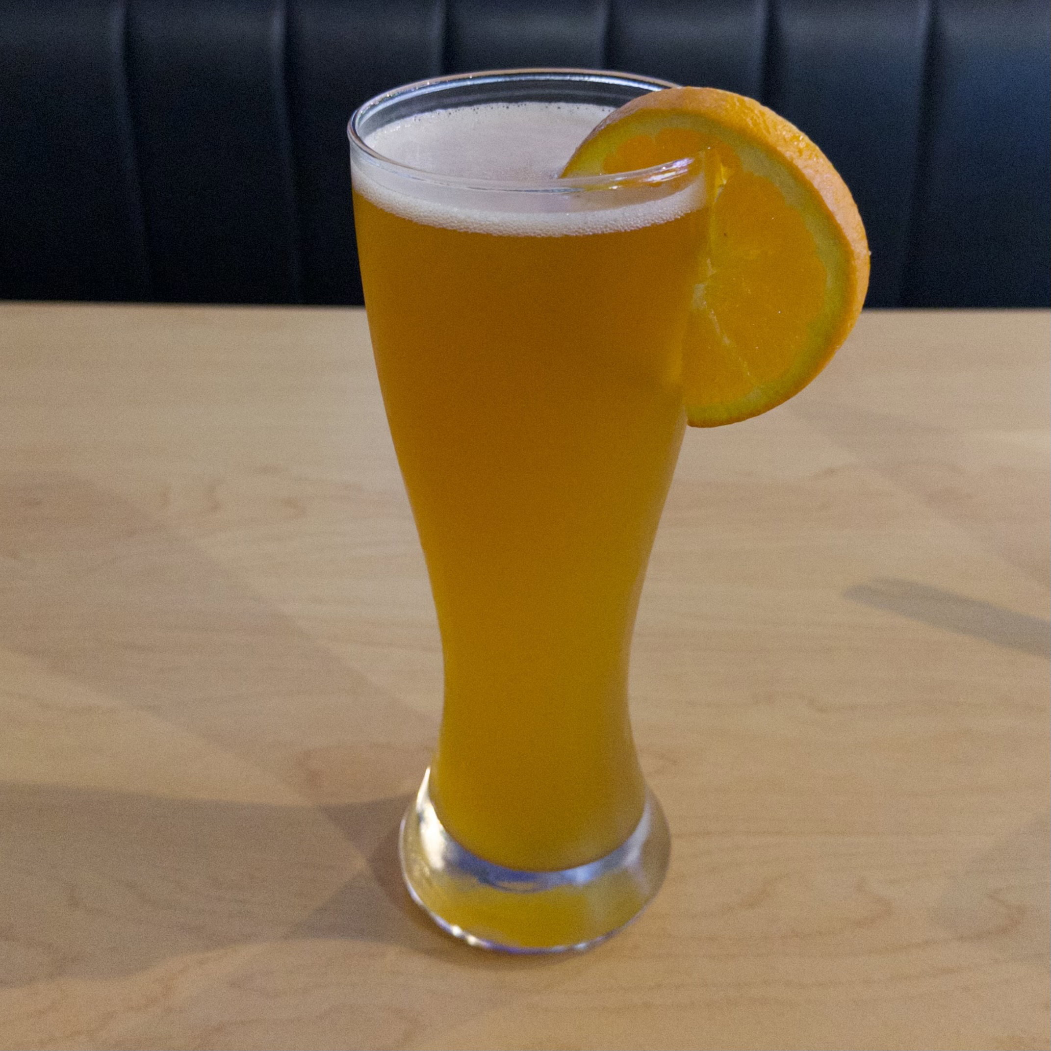 A tall glass filled with golden beer topped with white foam, garnished with an orange slice on the rim, sitting on a light wooden surface against a dark quilted background.