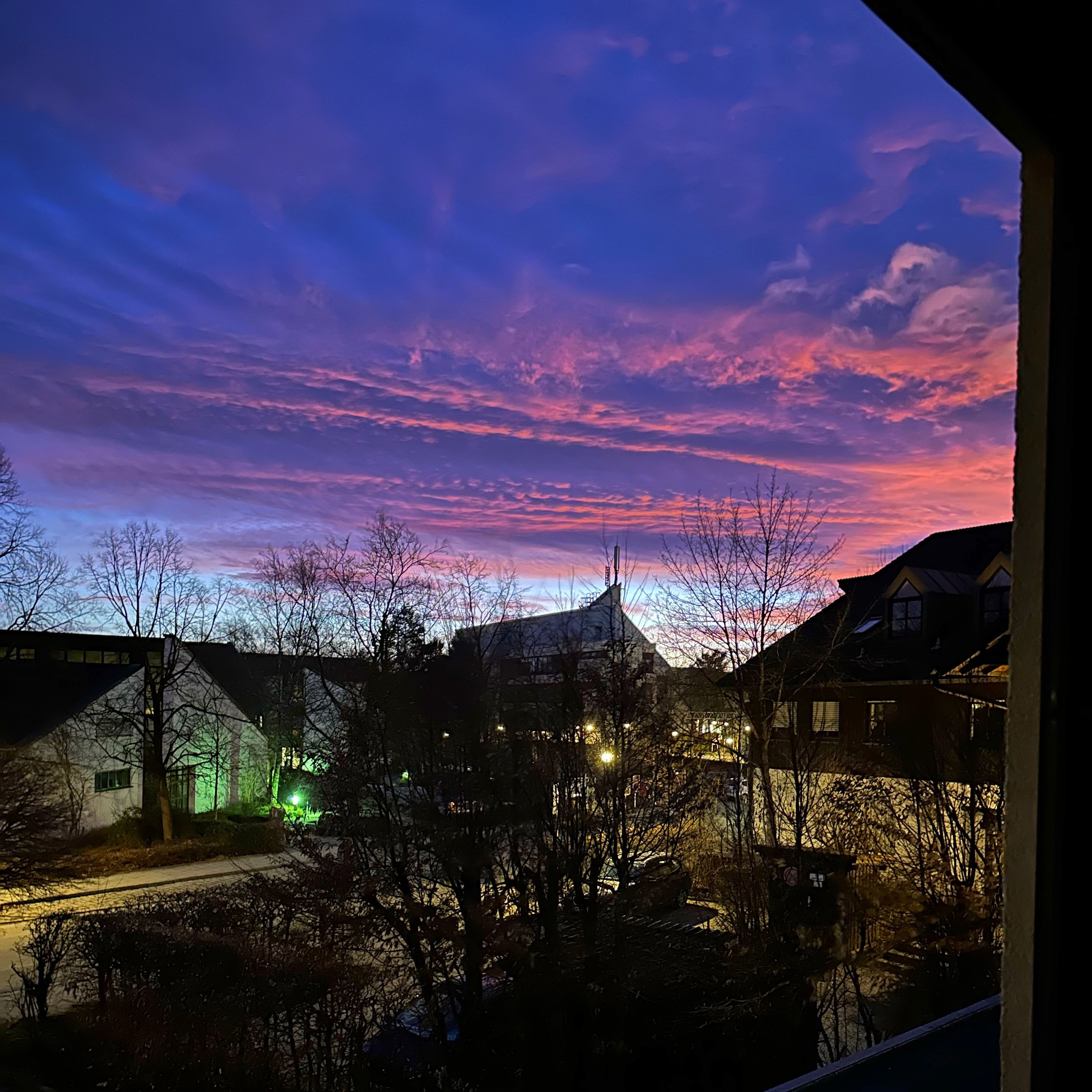 The sun is already set, but is still illuminating the purple cloud with rose streaks. The sky above the horizon is turquoise.  The view is out the first story of a building looking on to street, some buildings and trees are around 