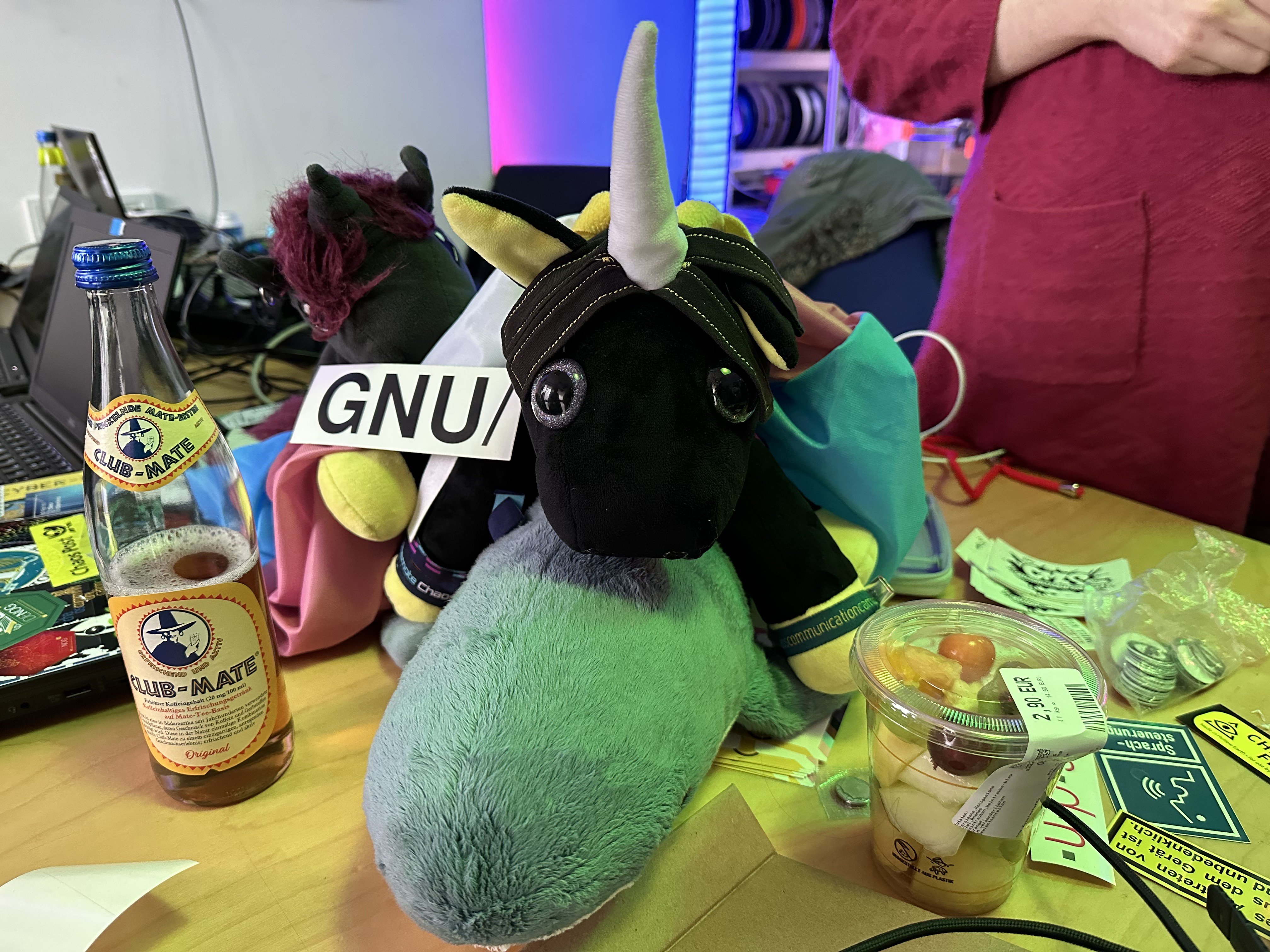 A GNU/Sticker next to Ida (black unicorn with yellow accents) on a smolhaj. There is a bottle of mate and some sticker around, another Unicorn and a transflag in the background of the pile