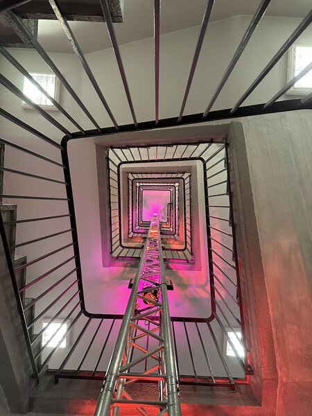 Looking straight up a square winding staircase with metal railings going round.
There is a truss attached to the railings all the way up to the 5th floor. It's used as a mounting point for several pink and orange lights colouring the ceiling of the floors