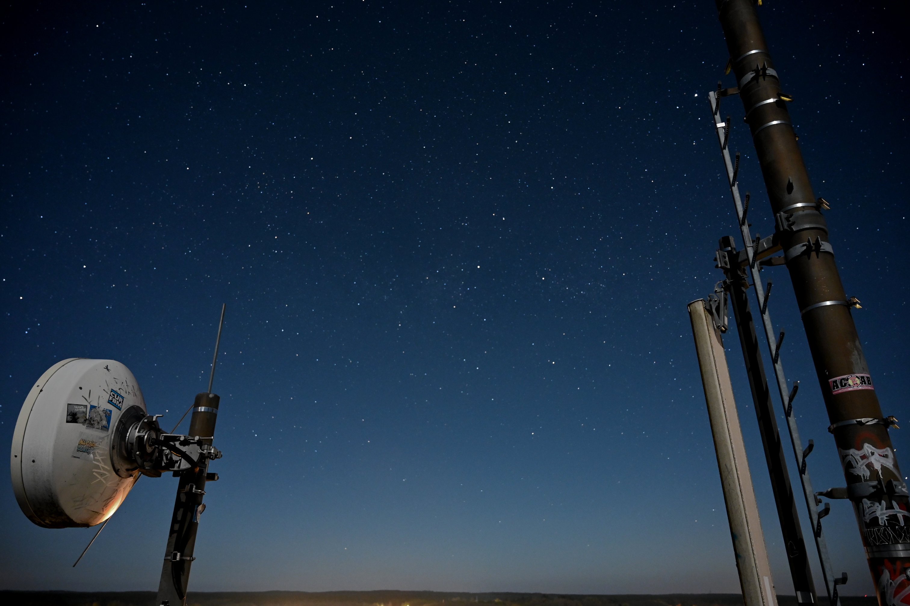 A Picture of the night's sky.  It's framed by the horizon from below, going from a warmish white with barely any stars visible to a ver deep blue with lots of stars.  on the left there is a directional radio antenna with some stickers on it. On the right is a mobile phone pole with ladder and antenna