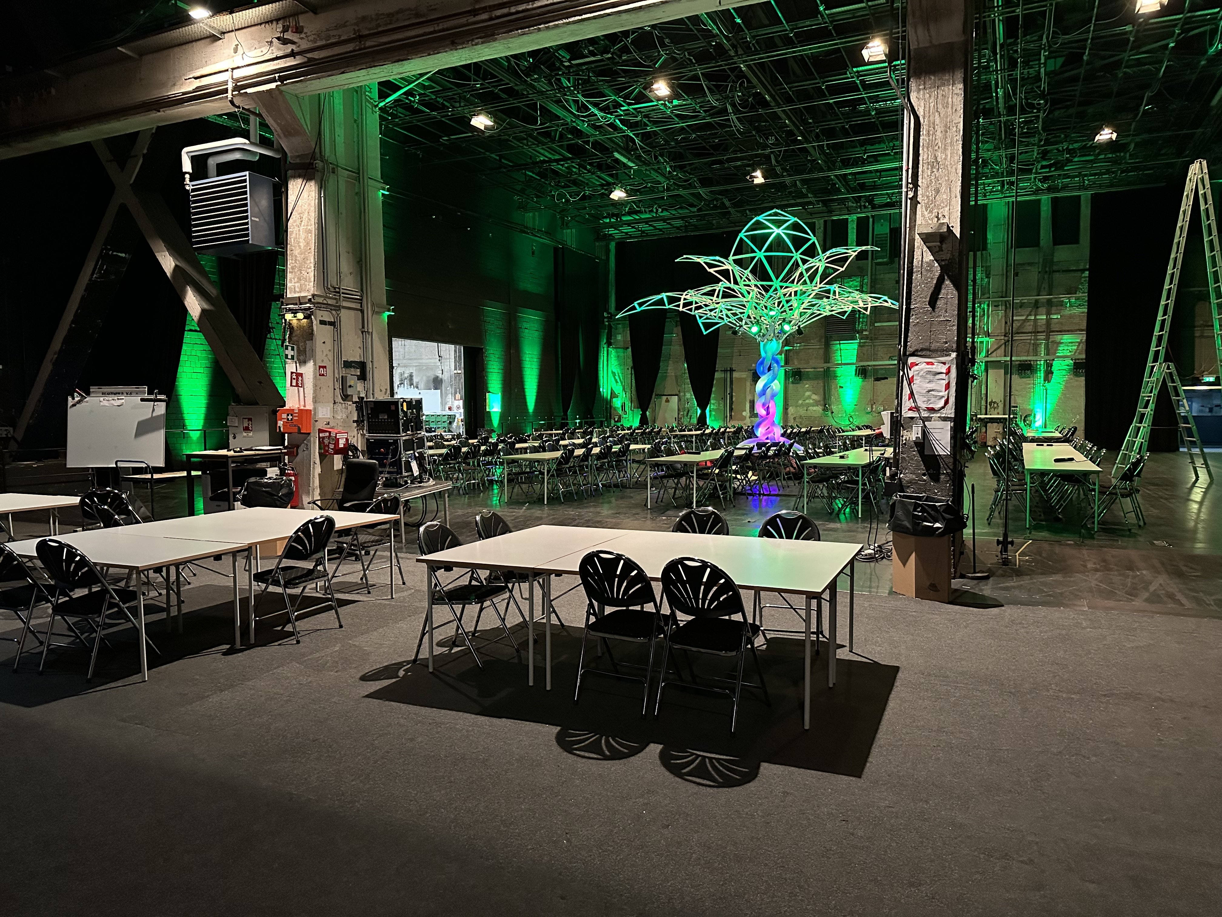 The main Easterhegg 2023 hackcenter. The evening before the event, empty.  A hall filled with desks and chairs in groups and rows. There is colourful green lighting and an RGB fake palm tree in the middle. The main lighting is still on.