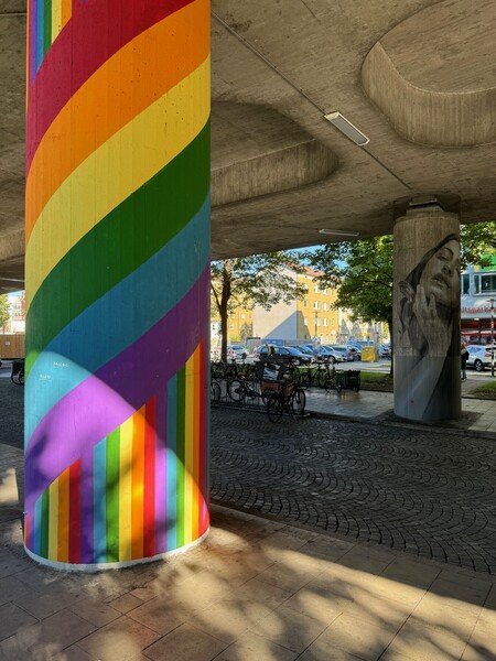 Under a concrete bridge, one stud is painted in rainbow colours the other one has a drawing of a woman on it