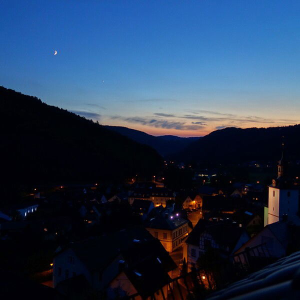 The image shows the village of Muggendorf during dusk. The sky is a deepening blue with the last hints of orange and pink from the setting sun on the horizon. A crescent moon is visible in the sky, adding a serene touch. The village is nestled in a valley surrounded by dark, forested hills. Streetlights and windows illuminate the buildings, casting a warm, cozy glow throughout the village. A church with a prominent steeple stands out in the scene, enhancing the quaint and peaceful atmosphere of this picturesque village.