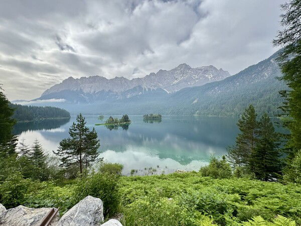 The picture shows the Wetterstein mountain range towering over Lake Eibsee. It is early summer, and the plants and trees in the foreground are lush and vibrant shades of green. The weather is slightly hazy, but the cloud cover is beginning to break, allowing patches of sky to peek through. The lake is calm, reflecting the surrounding landscape and the towering mountains in its serene waters. The overall scene is tranquil and picturesque, capturing the natural beauty of the area.