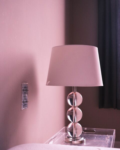 The picture shows a stylish table lamp placed on a transparent side table. The lamp has a modern design, featuring a metallic base with three stacked, spherical glass elements supporting a simple, cylindrical lampshade. The background wall is painted in a soft pink hue, and there is a light switch panel mounted on the wall next to the lamp. Part of a dark curtain is visible on the right side of the image, suggesting the lamp is positioned near a window. The overall ambiance of the scene is calm and minimalistic.