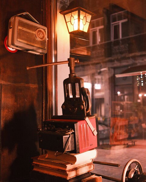The picture shows a cozy, vintage-themed interior scene. A small, lantern-style wall lamp emits a warm glow, illuminating the surrounding area. Mounted on the wall is a retro portable radio. Below the lamp and radio, there is an old-fashioned metal vice clamp placed atop a stack of old books and vintage electronic equipment, including a red device with dials and wires. The setting appears to be near a window, with reflections of a street and buildings outside, adding to the nostalgic ambiance of the scene. The overall atmosphere is warm and inviting, with a touch of rustic charm.