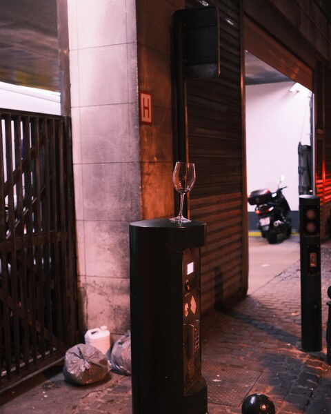 The picture shows an urban street scene at night. In the foreground, there is a black bollard with an empty wine glass placed on top of it. The bollard is positioned near a stone wall with a metal gate and a rolling shutter door leading to a dimly lit garage or alleyway. A scooter is parked inside the garage. Nearby, a few garbage bags and a white container are placed against the wall. The scene is illuminated by artificial streetlights, casting a warm glow and creating shadows, giving the image a slightly gritty, urban feel.