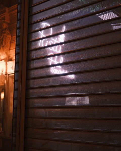 The picture shows the exterior of a storefront at night, viewed through a partially closed metal shutter. Behind the shutter, a neon sign glows with the words 'Pasta' and 'Sandwiches,' suggesting a restaurant or café. The sign emits a pinkish light that diffuses through the perforated metal, creating a soft, colorful glow. In the background, there are hints of the interior and some architectural details of the building next door, which is also illuminated by warm lights. The overall atmosphere is urban and slightly mysterious, with the interplay of light and shadows adding depth to the scene.