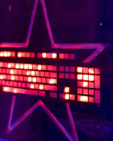 The picture shows a close-up of a brightly illuminated sign or display, featuring a star shape with lights arranged in a keyboard shape inside it. The lights emit a red glow, contrasting sharply against a dark background. 