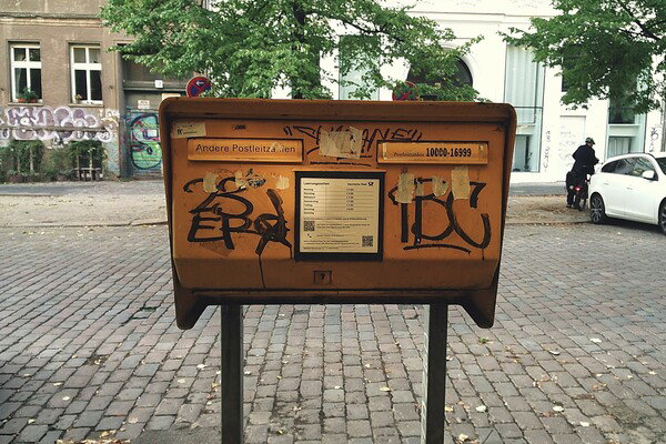 Graffiti-covered yellow mailbox on cobblestone street, old buildings, cyclist, parked car, and trees.