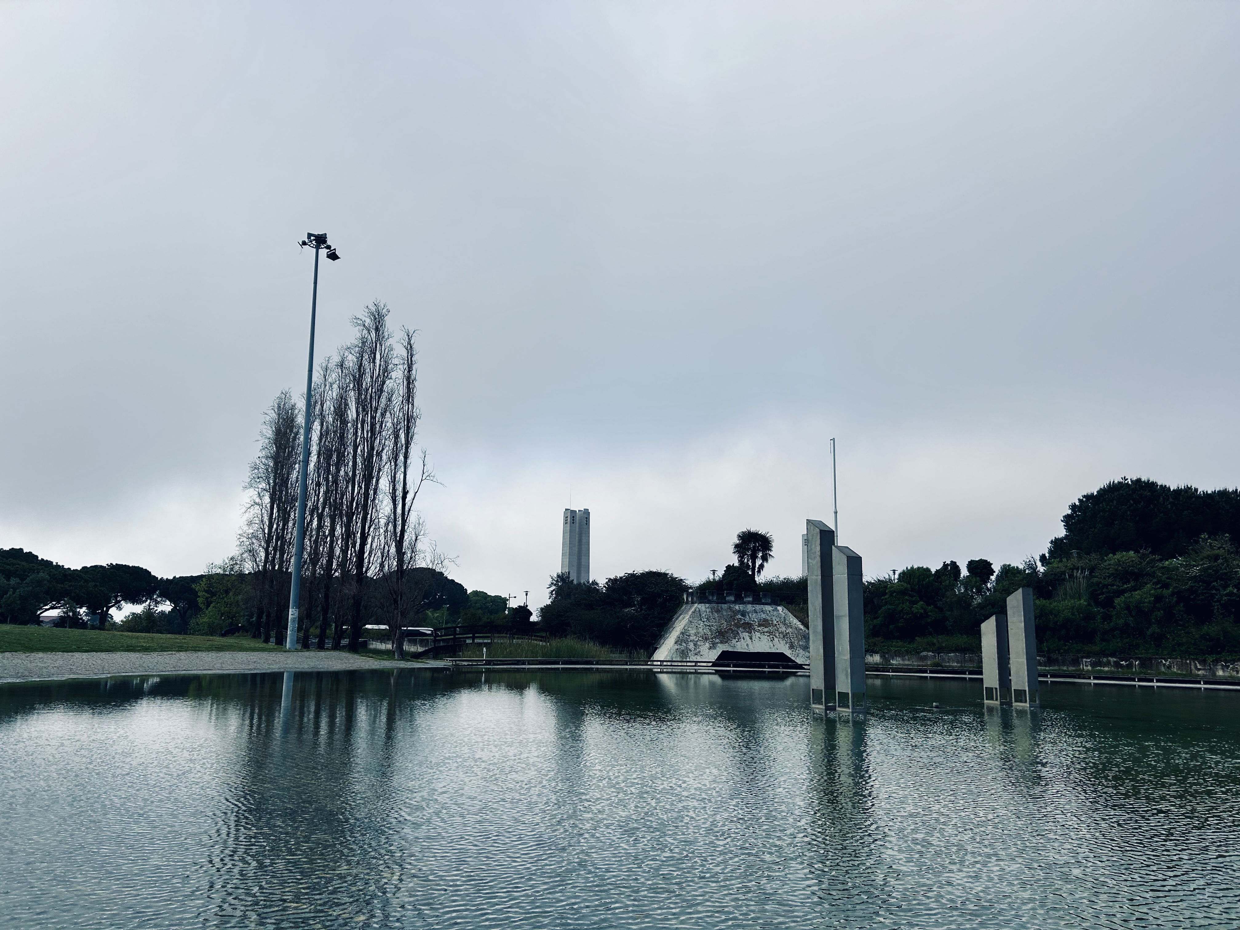 Pond with a couple of bevelled cement rectangles sticking out of the water - I'm not sure that they serve any practical purpose. Beyond it, a low treeline is visible, with a larger stone structure appearing in the distance.
