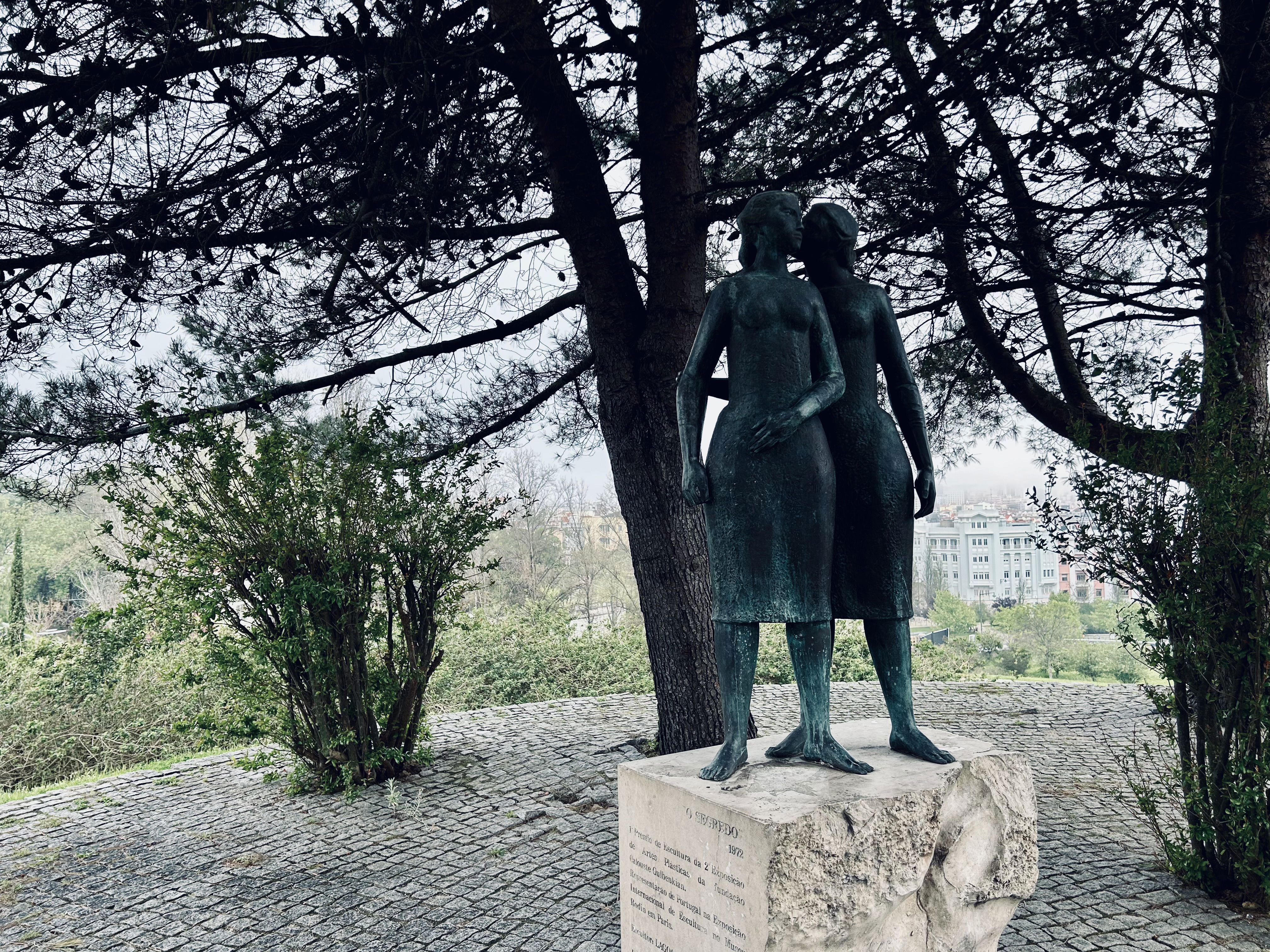 A statue of two girls standing close together. One is whispering in the other's ear while holding her arm. The statue is called "The Secret" and it stands on a circular plateau paved in stone. Around it are a couple of trees, and buildings are visible in the background.