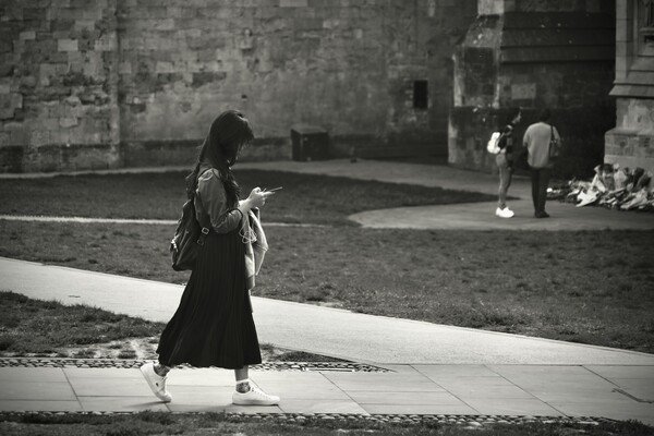 Monochrome street photography. A young woman with long skirt walks along the path in front of Exeter Cathedral, looking at her phone. In the background, people are laying flowers, out of focus - taken shortly after the Queen's death.