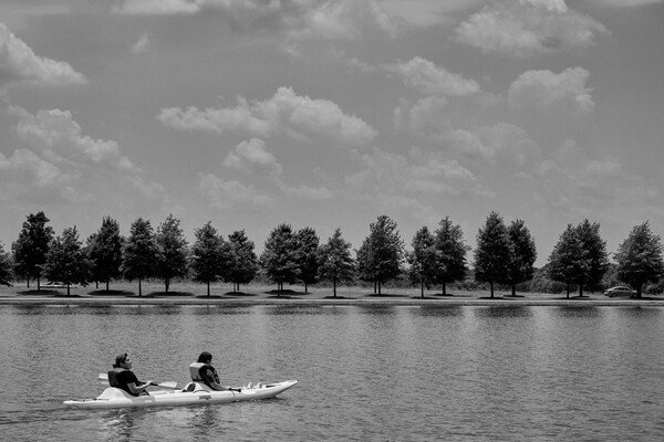 A black and white photo of two people in a kayak on a lake. There is a row of trees on the opposite bank, and clouds in the sky.