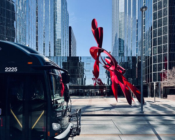 A glass building with a modern red art installation in front of it. A black bus is entering the photo from the left passing through.