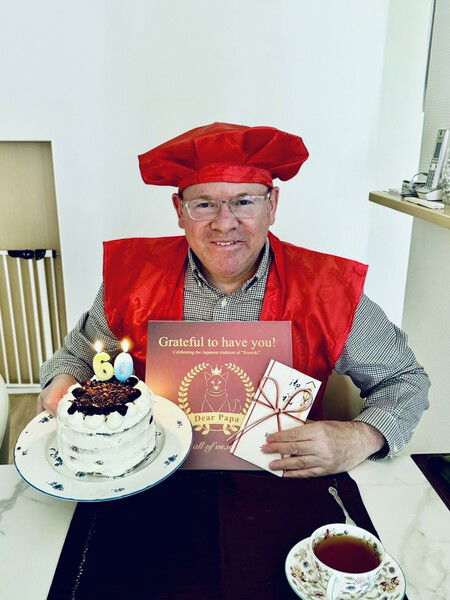 Man in a red chef's hat holds a candlelit cake with a table and celebratory decorations.