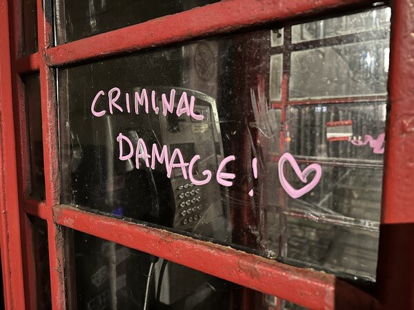 A phone box window with graffiti in pink that says Criminal Damage! and a little heart next to it.