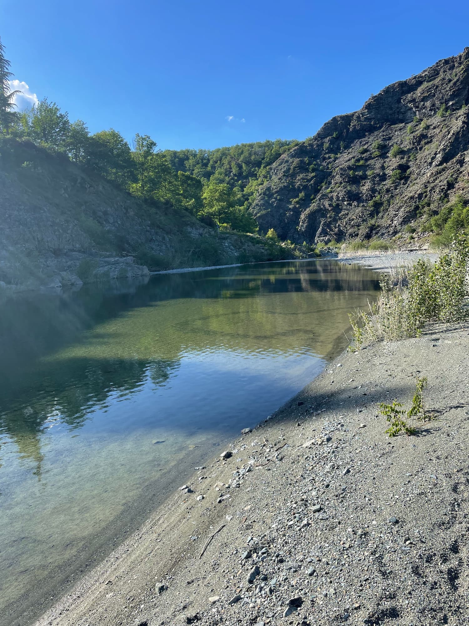 A picture of a river, with low water and a blue, bright sky. There is a small beach. On the right, a rocky and steep edge. On the horizon, there are green trees on a hill.