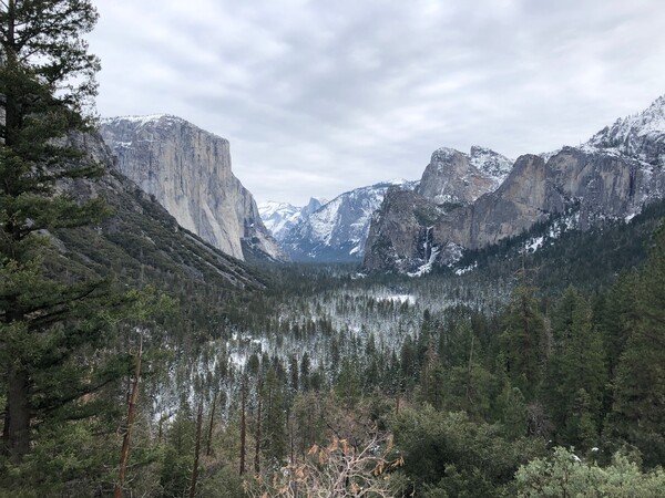 The view from the Tunnel View scenic location at Yosemite National Park in California, with trees near the valley, and mountains at a distance, and 2 large mountains close by (El Capitan on the left, and Bridalveil Falls on the right)