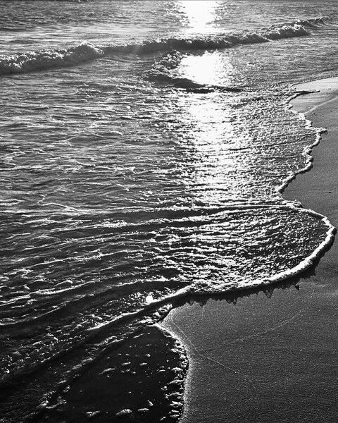 Black and white photo of waves lapping the shore at sunset in Gulf Shores, Alabama along the Gulf of Mexico. The sunlight is reflecting on the water at an angle.