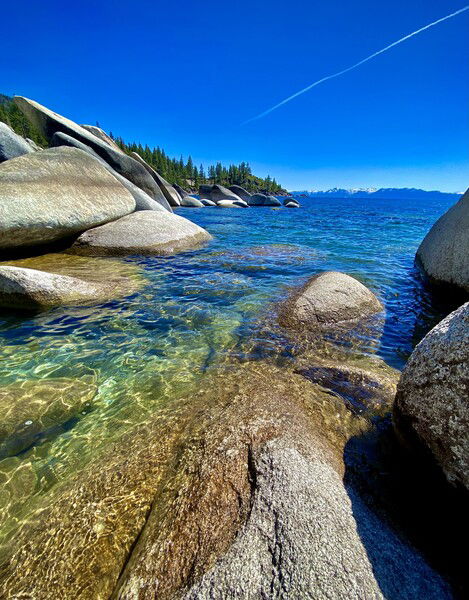 A sunny, blue sky day at Lake Tahoe, boulders line the shore, mountains in the distance. 