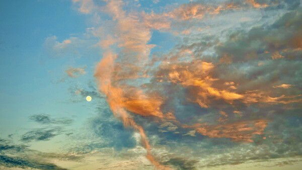 A nearly full moon peeks out from the colorful clouds at sunset