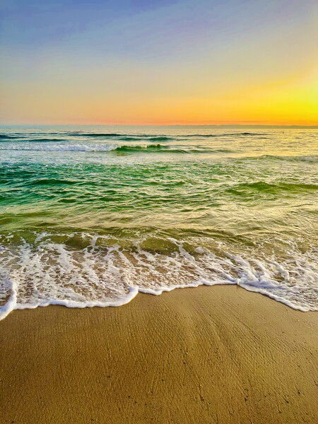 Gulf Shores, Alabama looking serene at sunset, featuring gentle waves lapping at a sandy shore, with a gradient sky transitioning from orange to blue.