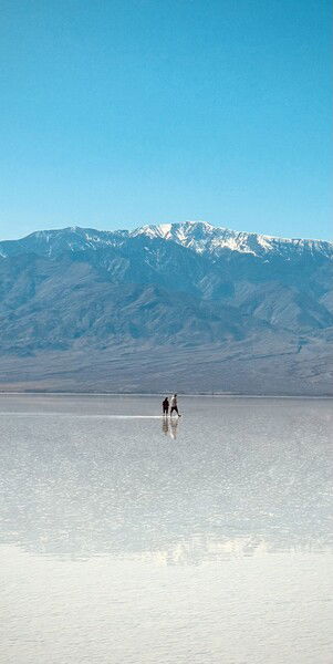 Two people walking on a shallow reflective lake formed on a salt flat with a backdrop of snow-capped mountains under a clear blue sky at Death Valley
